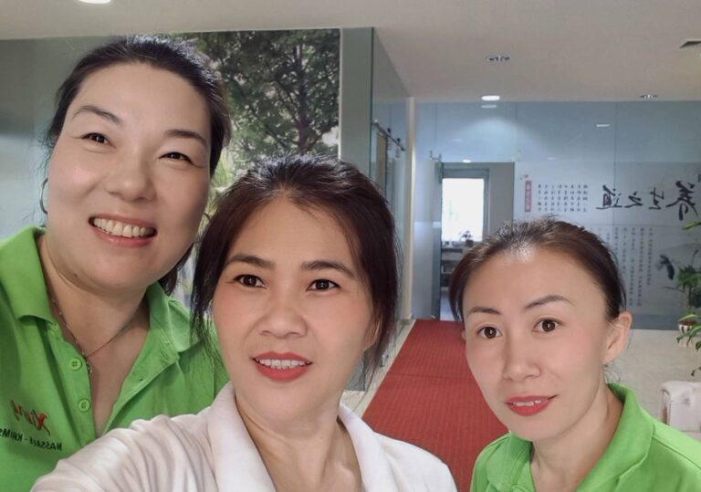 Drei lächelnde Masseurinnen in einem eigenen Massagestudio, grüne und weiße Shirts, Gruppenfoto bis zur Schulterhöhe, professionelle Atmosphäre für Wellness und Entspannung.