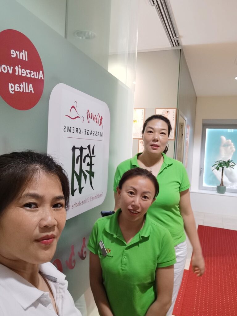 Drei lächelnde Masseurinnen in einem eigenen Massagestudio, grüne und weiße Shirts, Selfie, professionelle Atmosphäre für Wellness und Entspannung.