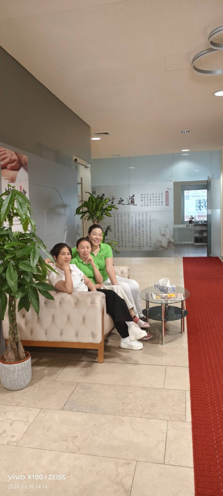 Drei lächelnde Masseurinnen sitzen in ihrem Massagestudio auf einer hellen Couch, grüne und weiße Shirts, vor ihnen ein kleiner Glastisch, umringt von zwei gut gepflegten Pflanzen, professionelle und beruhigende Atmosphäre für Wellness und Entspannung.