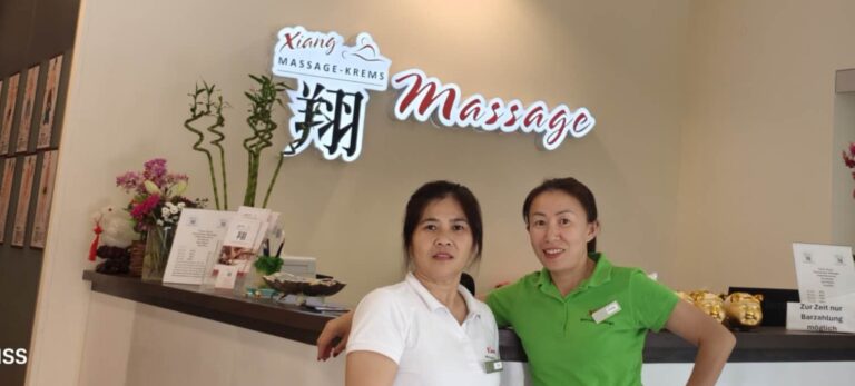 Zwei freundliche Masseurinnen in einem eigenen Massagestudio, grünes und weißes Shirt, im Hintergrund der Empfang und das Logo von Xiang Massage in Rot, Weiß und Schwarz